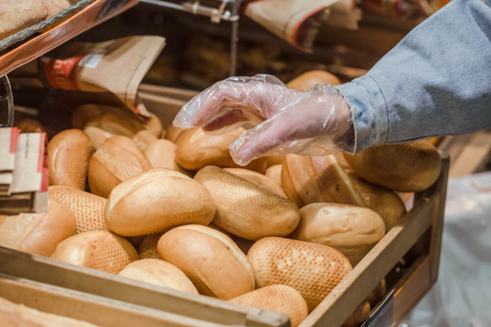 A person wearing a plastic glove selects fresh bread rolls from a wooden display at a bakery.