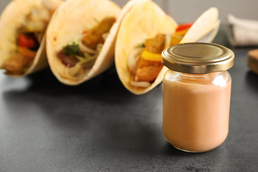 A jar of creamy fish taco sauce in the foreground with three loaded fish tacos filled with colorful vegetables in the background on a dark surface.