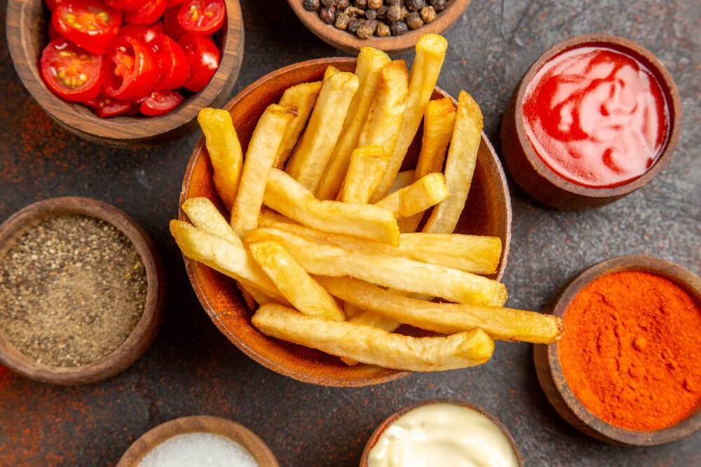 A bowl of golden French fries surrounded by various condiments and spices, including ketchup, mayonnaise, black pepper, chili powder, and sliced cherry tomatoes.