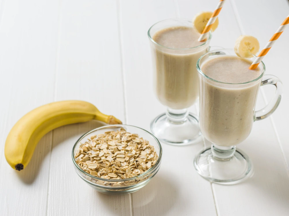 "Two glasses of creamy banana oatmeal smoothie with striped straws, accompanied by a banana and a bowl of rolled oats on a white wooden table."