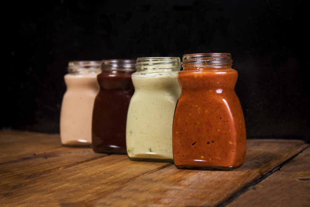 Four glass jars of different sauces arranged on a rustic wooden surface, featuring creamy pink, rich chocolate brown, green, and vibrant red sauces against a dark background.