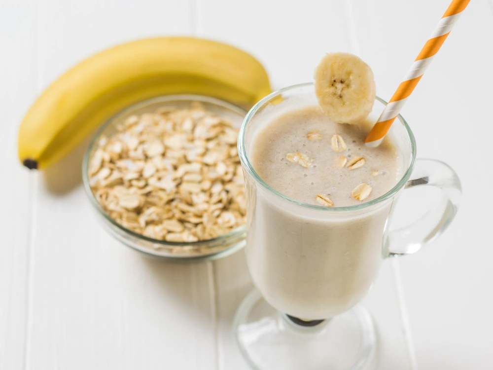 "A single glass of banana oatmeal smoothie garnished with a banana slice and striped straw, placed next to a fresh banana and a bowl of rolled oats on a white wooden table."