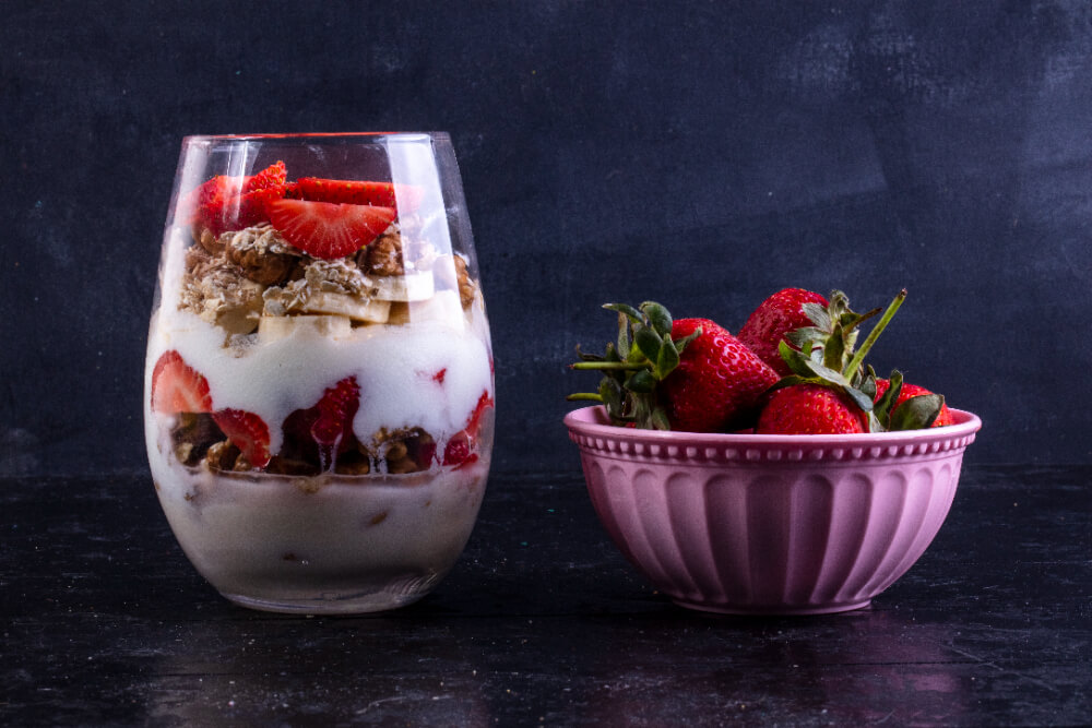 A delicious yogurt parfait in a clear glass, layered with fresh strawberries, granola, and banana slices, accompanied by a pink bowl filled with ripe strawberries