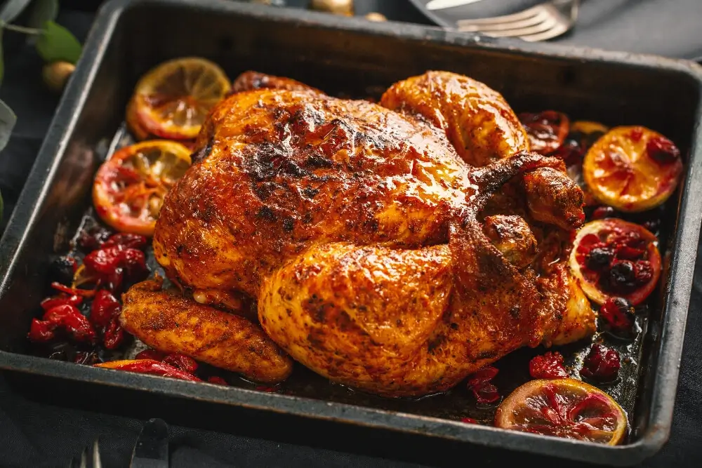 Golden slow roasted chicken with a crispy skin, surrounded by roasted citrus slices and cranberries in a black roasting pan.