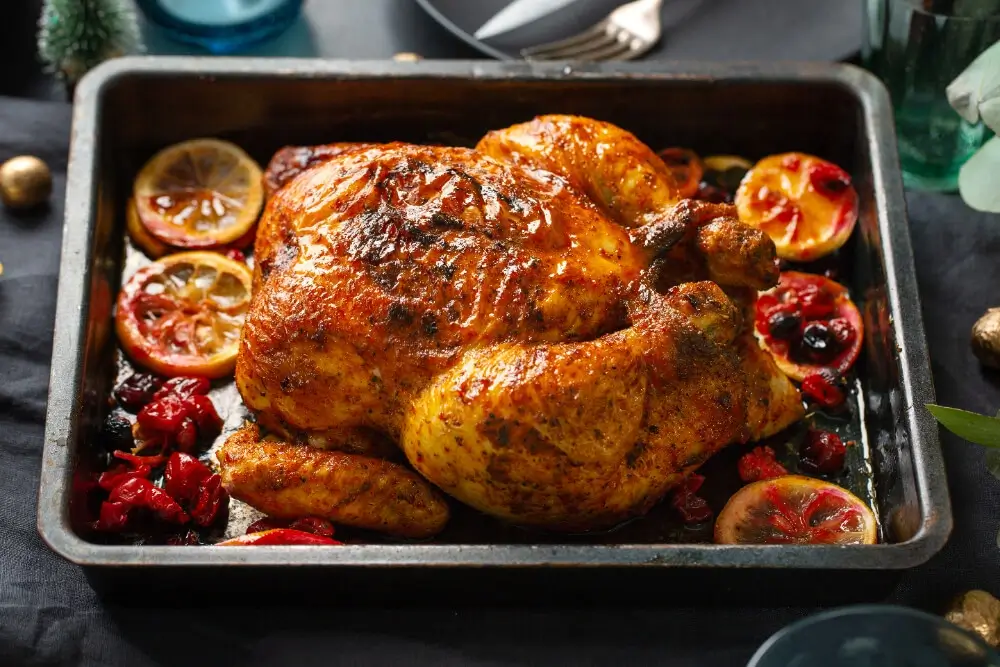 Golden-brown slow roasted chicken in a roasting pan, surrounded by citrus slices and cranberries, set on a festive table.