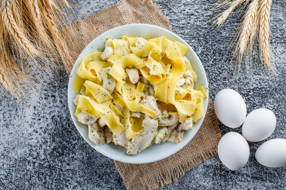 Top view of chicken spaghetti carbonara with wide pasta ribbons on a rustic surface, surrounded by wheat and fresh eggs.