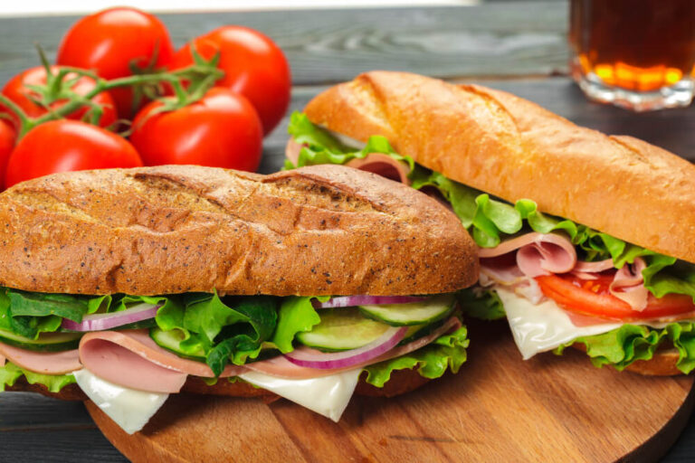 Two freshly made grinder sandwiches with lettuce, tomatoes, cucumber, and deli meats served on a wooden board, with ripe tomatoes in the background.