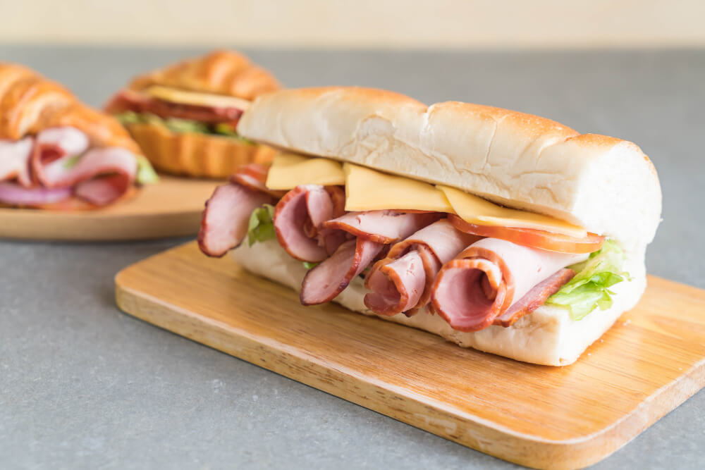 A freshly prepared grinder sandwich with layers of deli meats, cheese, lettuce, and tomatoes on a soft roll, served on a wooden board.