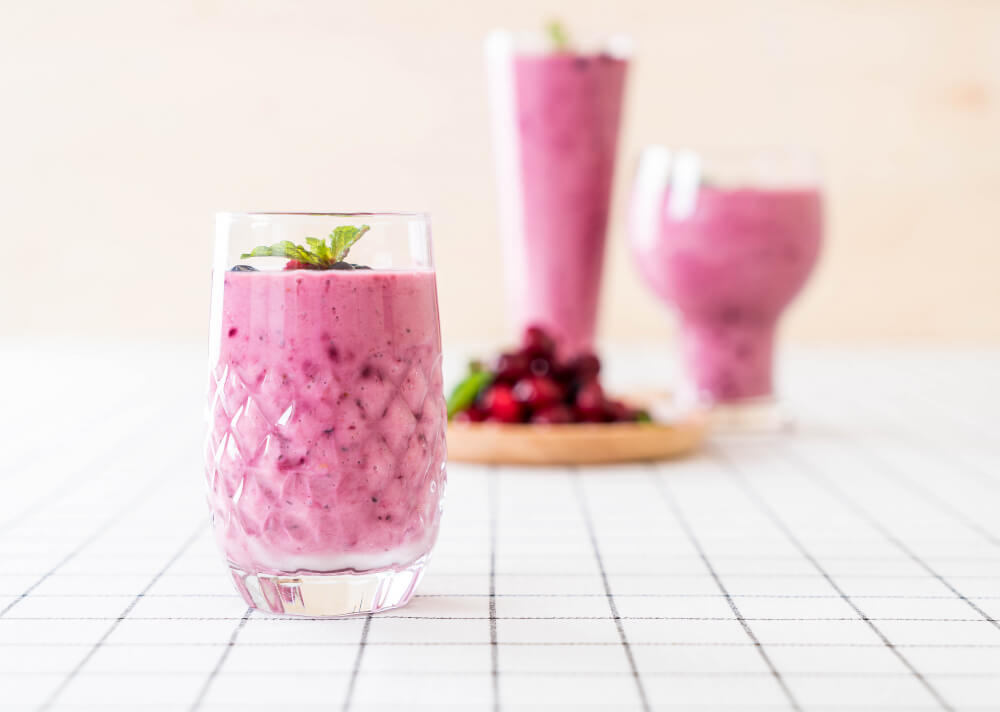 Three berry smoothies in elegant glasses, with the front glass garnished with mint leaves and blueberries, set on a white checkered table.