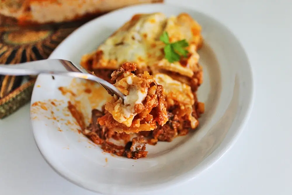 A close-up of a serving of crockpot lasagna on a white plate, with a fork lifting a cheesy, meaty bite.