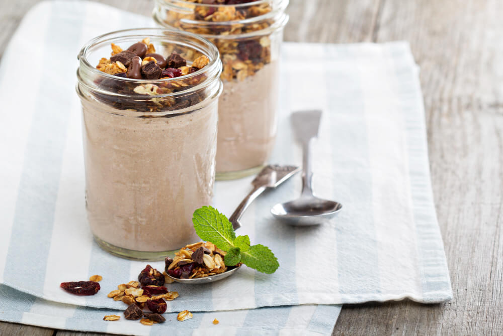 Two mason jars filled with creamy chocolate overnight oats, topped with granola, chocolate chips, and cranberries, placed on a striped cloth napkin with spoons and scattered toppings nearby.