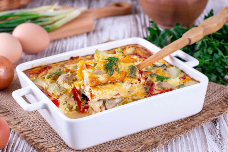 A freshly baked egg and vegetable casserole with a wooden spatula lifting a slice, garnished with fresh herbs in a rustic setting.