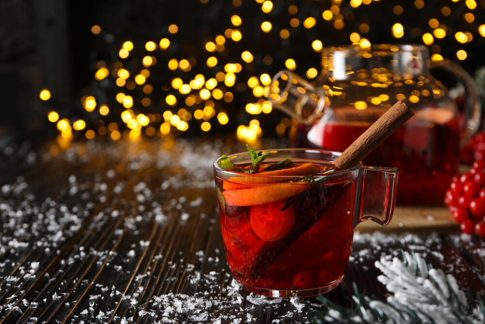 A glass mug of non-alcoholic Christmas punch with cinnamon sticks, orange slices, and cranberries on a snowy wooden surface with festive lights in the background.