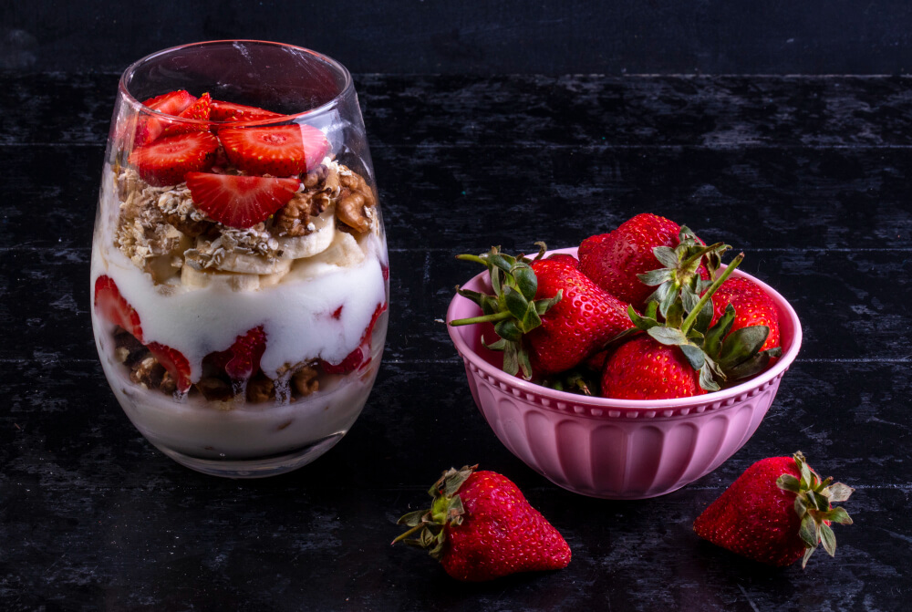 A layered yogurt parfait in a clear glass with fresh strawberries, bananas, granola, and walnuts, paired with a pink bowl of ripe strawberries on a dark textured background.