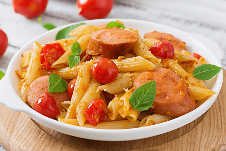 A plate of penne pasta with sliced sausage, cherry tomatoes, and fresh basil leaves.